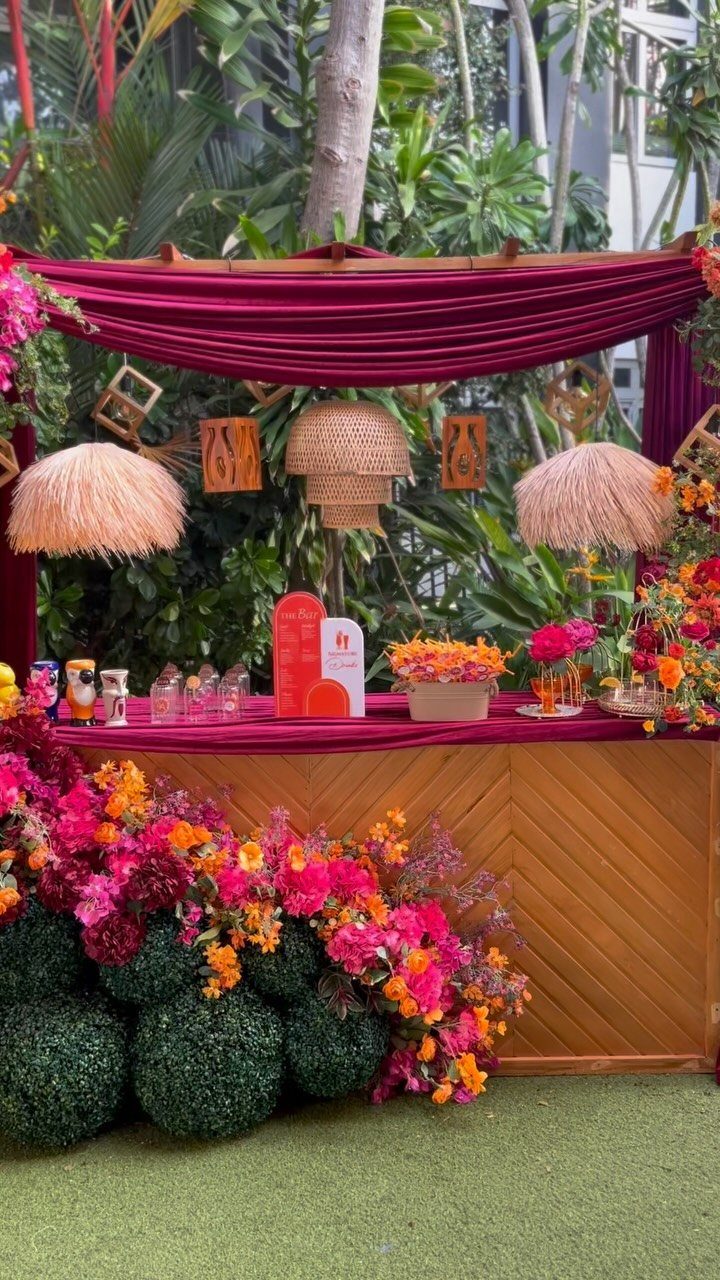 Wedding Decor and Themes: 12 Ideas From Beach to Garden 5 A decorated outdoor bar with magenta fabric, woven lamps, and colorful floral arrangements against tropical greenery.