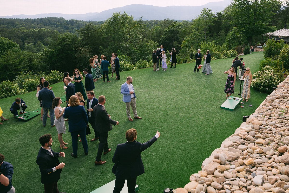 Guests playing cornhole on a green lawn during an outdoor wedding reception with mountains.