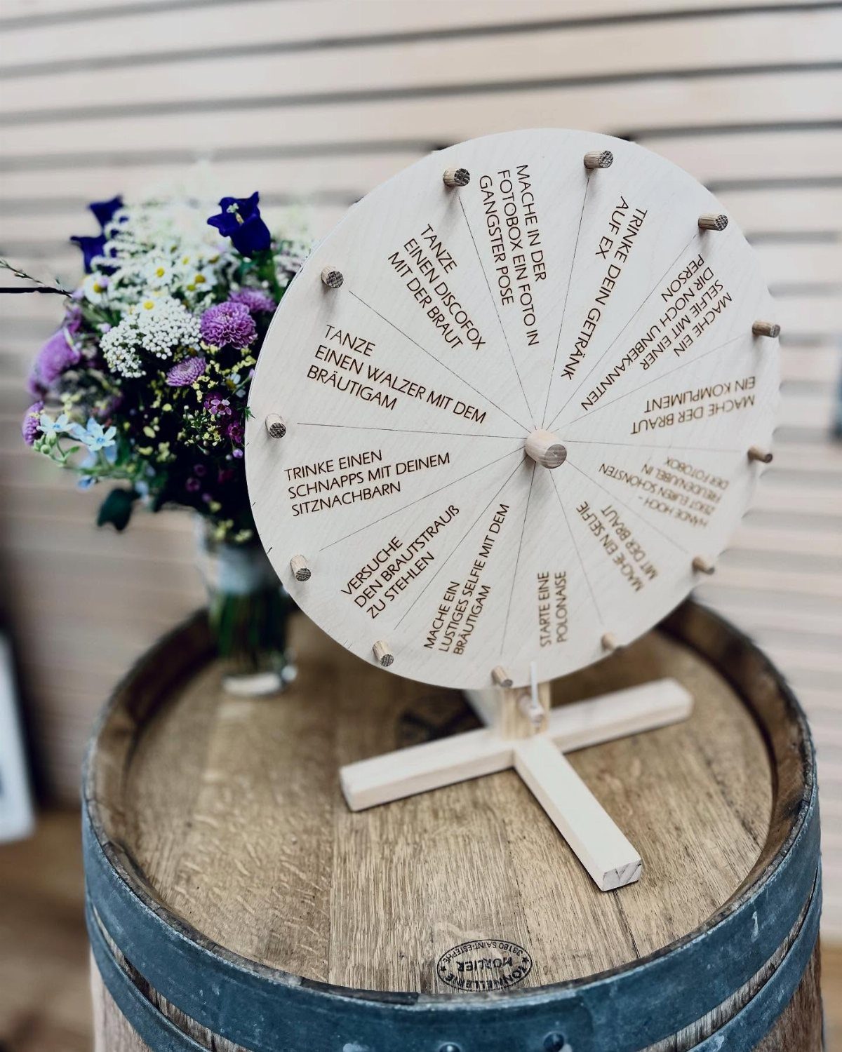 A wooden spinning prize wheel with German text sitting on a rustic barrel next to flowers.