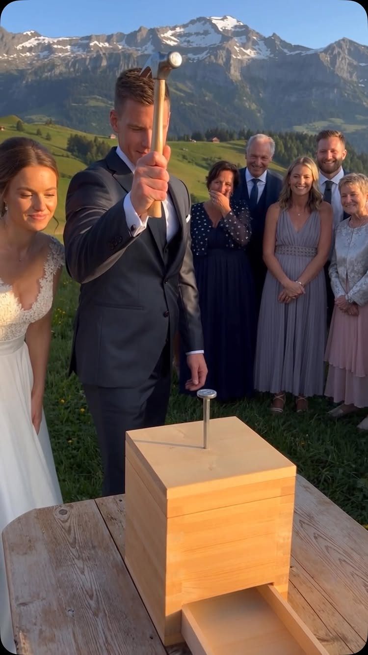 A groom uses a wooden hammer to hit a nail on a wooden box during a wedding reception.
