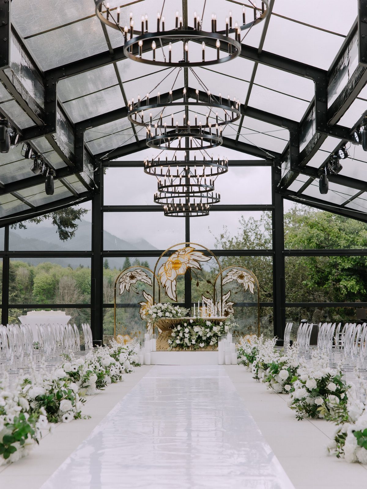 A wedding ceremony setup inside a glass pavilion with white flowers and clear chairs.
