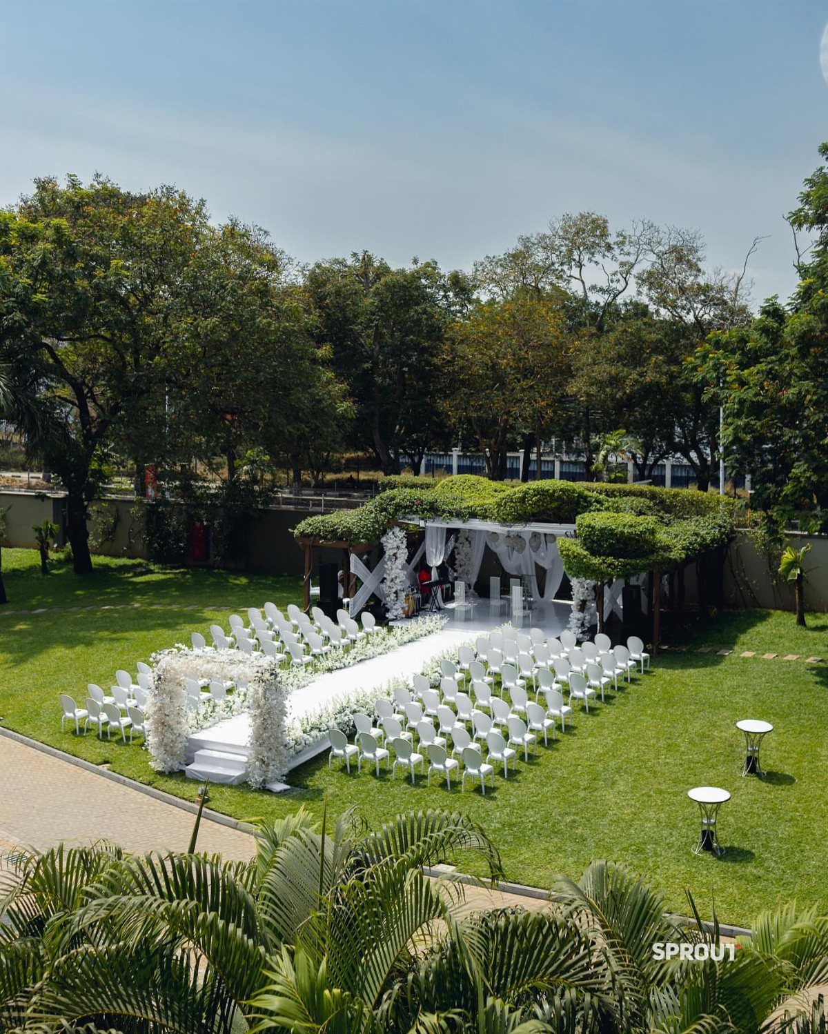 White wedding chairs and floral altar arranged on a green lawn in an intimate garden.