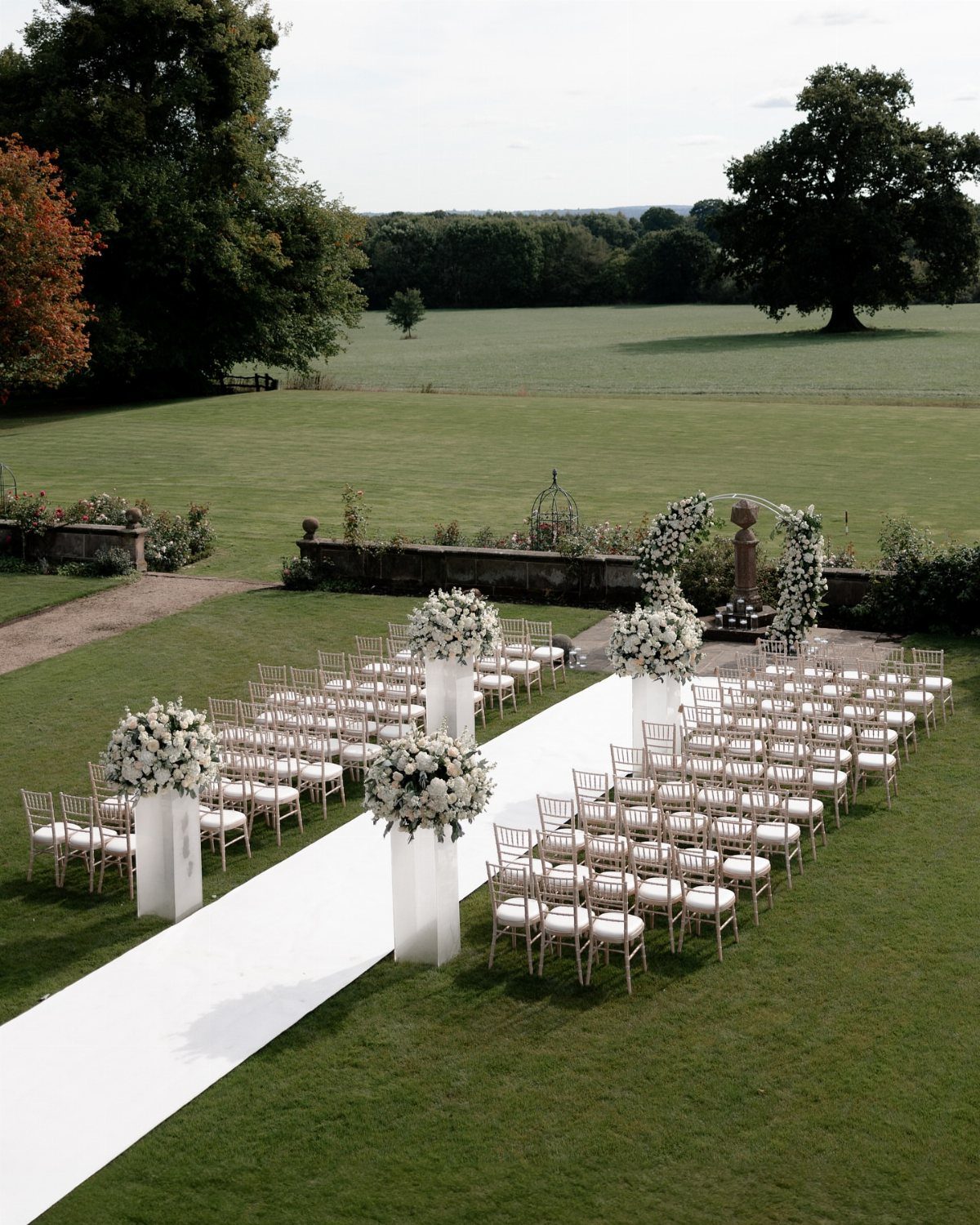 An outdoor wedding ceremony setup with white chairs, a white aisle runner, and floral arrangements on a lawn.