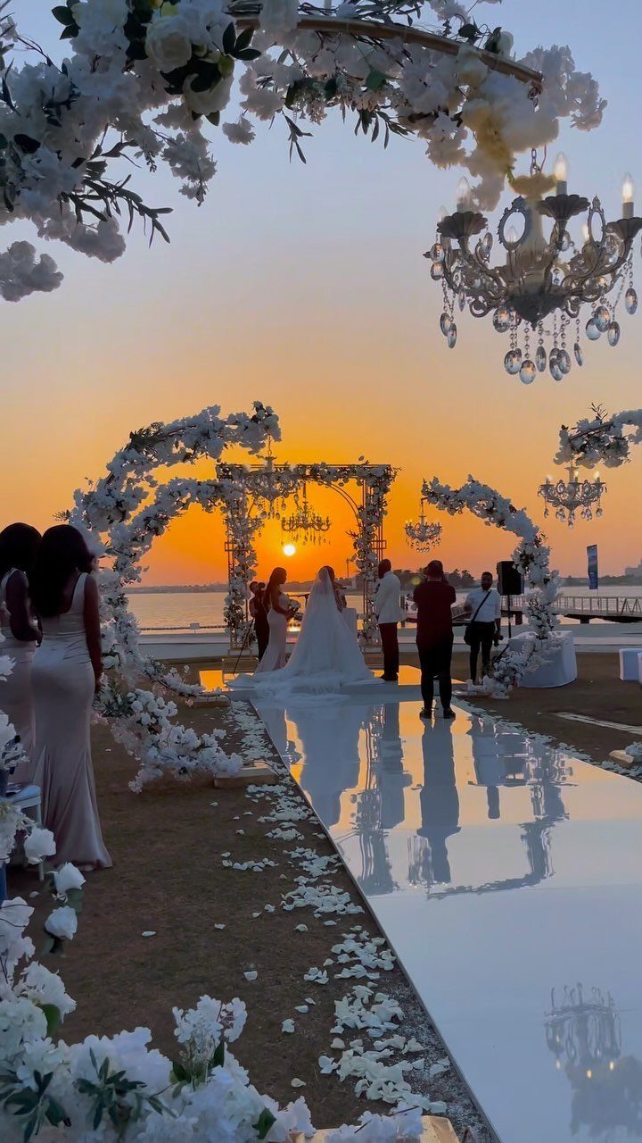 Wedding ceremony setup with white floral arches and chandeliers overlooking the ocean at sunset.