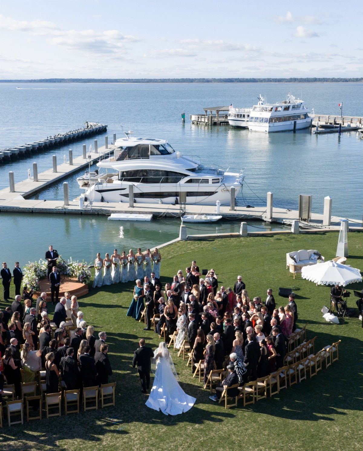 A wedding ceremony is held on a grassy lawn next to a marina with boats.