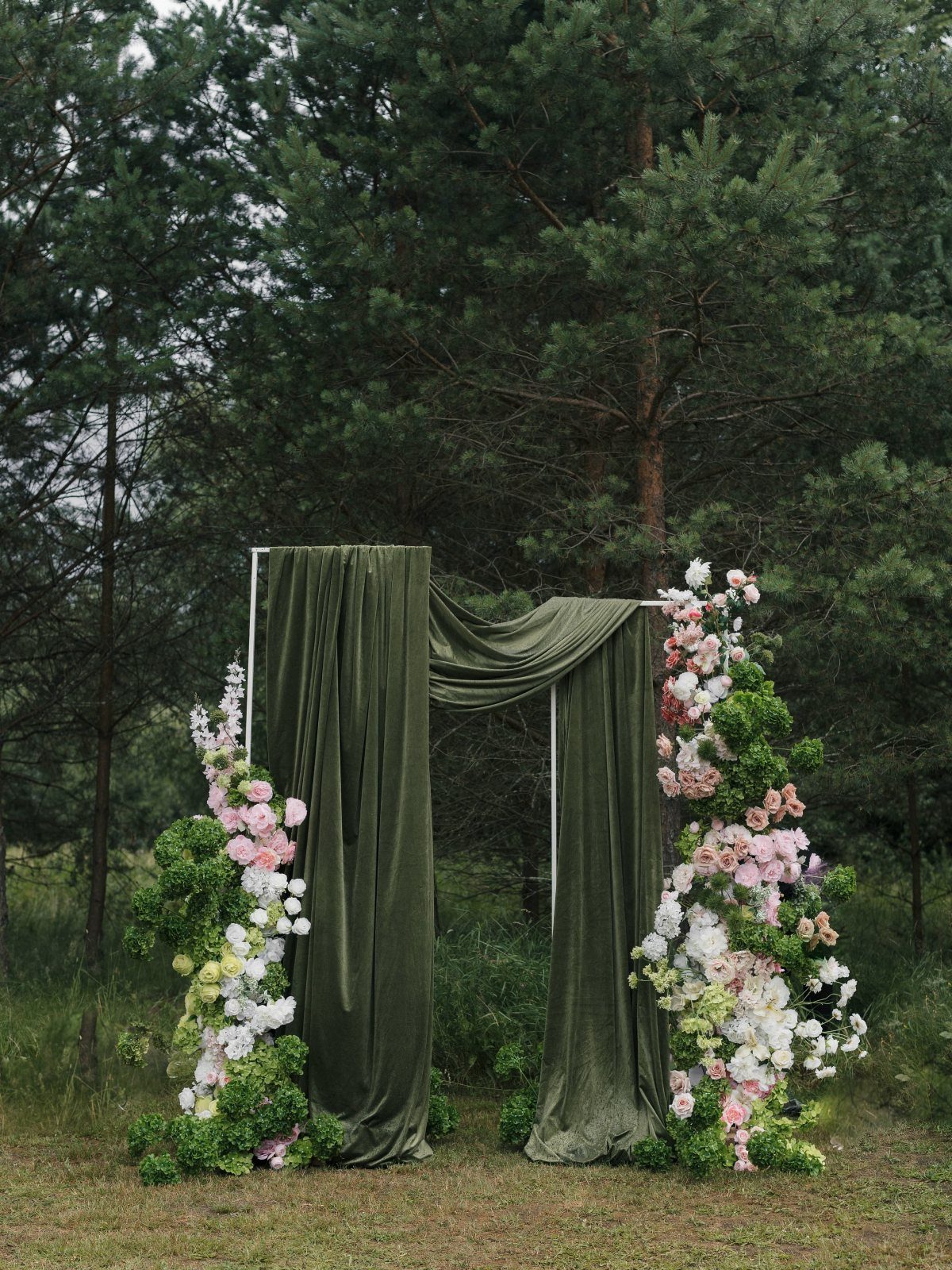 A wedding arch draped in green fabric and decorated with pink and white flowers in a forest.