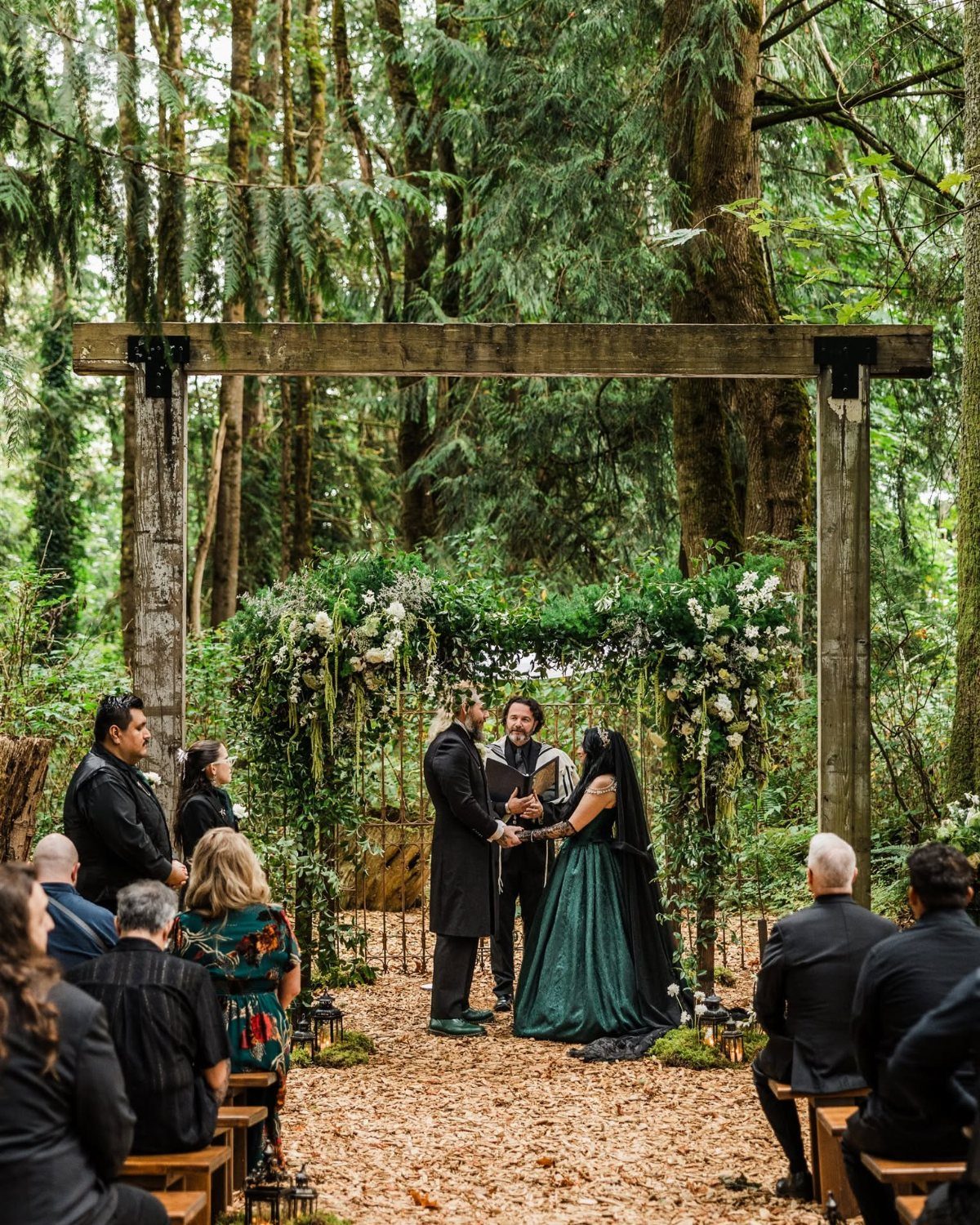 A wedding ceremony held in a forest under a floral wooden archway with guests seated.