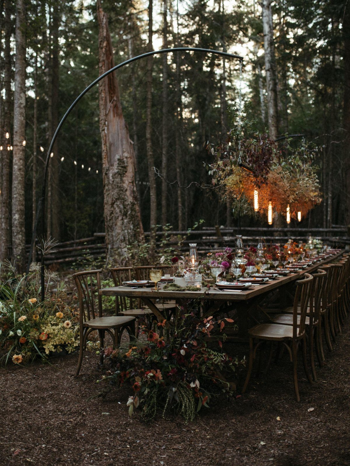 A long wooden reception table set in a forest under a large arch of hanging greenery and candles.