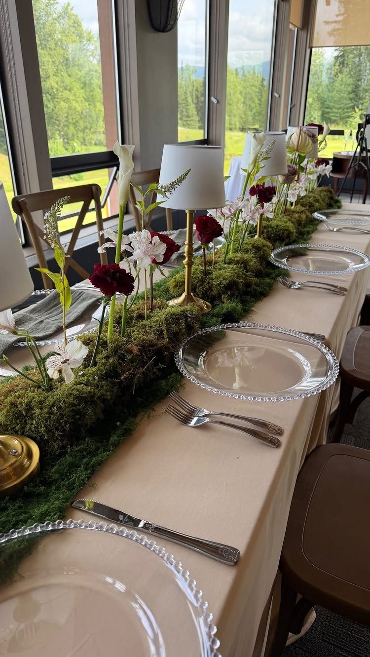 A long dining table featuring a thick green moss runner decorated with red and white flowers.