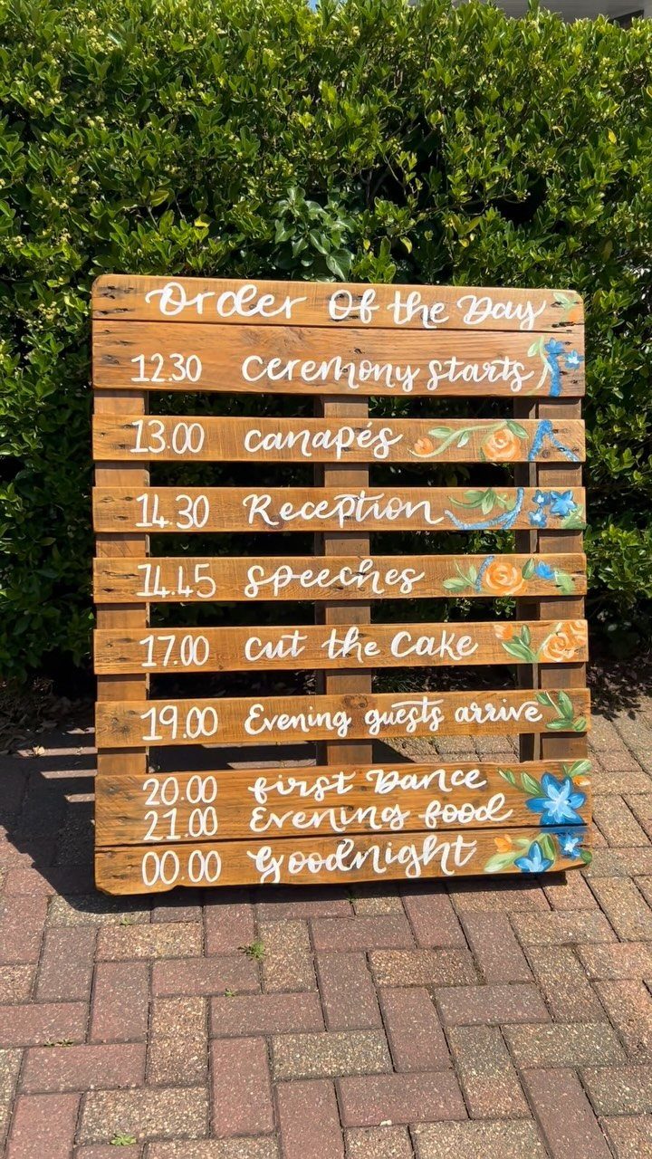 A wooden wedding schedule sign with floral decorations standing on a brick walkway near green bushes.