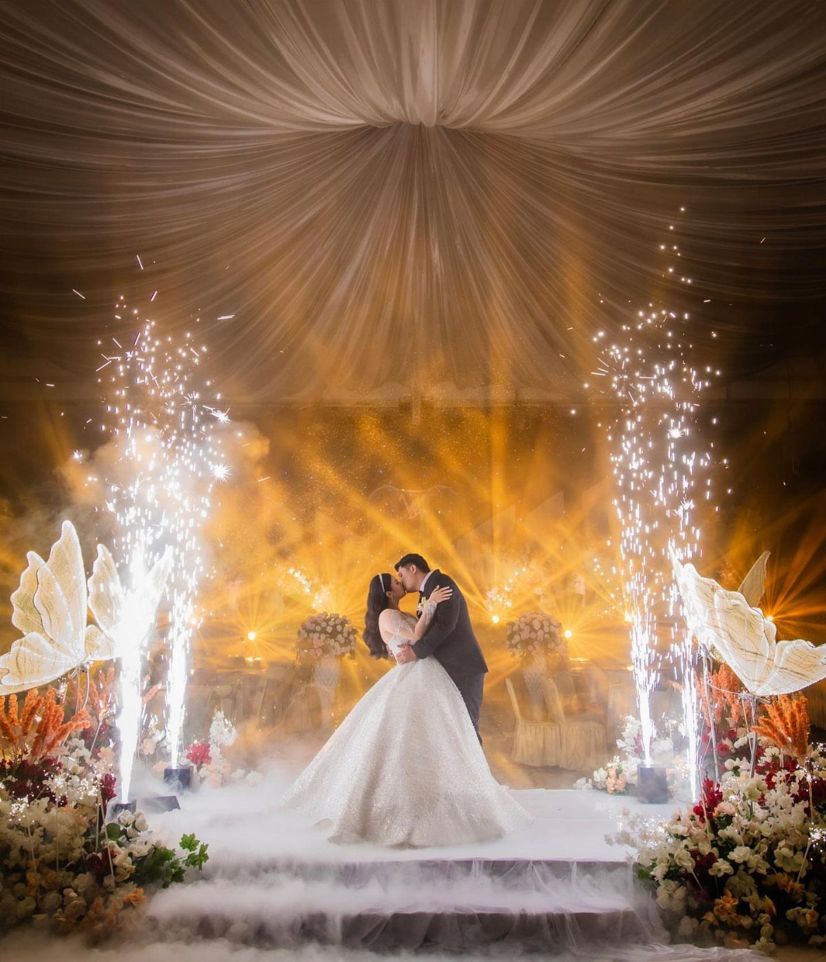 A bride and groom kissing on a stage surrounded by sparklers and fog.