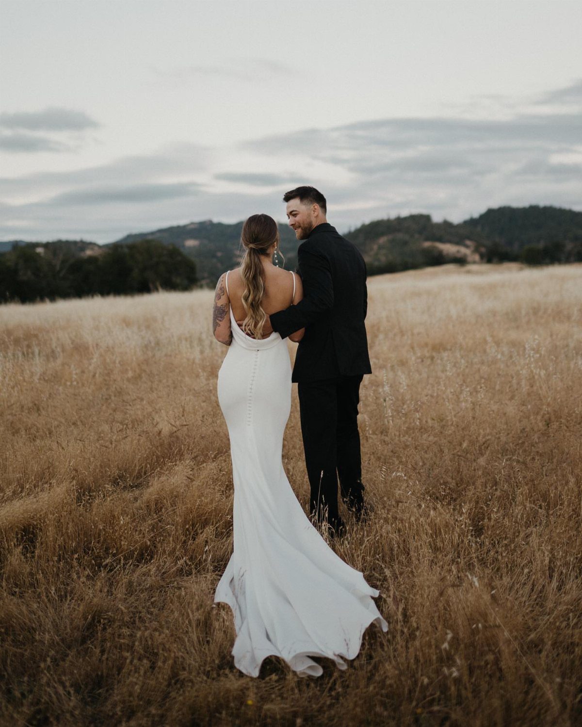 A bride and groom walking through a golden field while looking at each other.