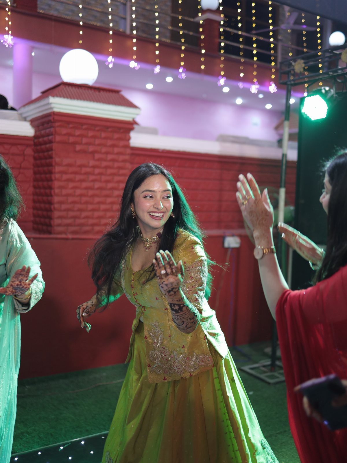 A woman in a green lehenga dancing joyfully at an outdoor evening wedding celebration.