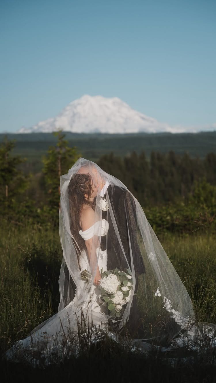 A bride and groom embracing in a field with a mountain in the background.