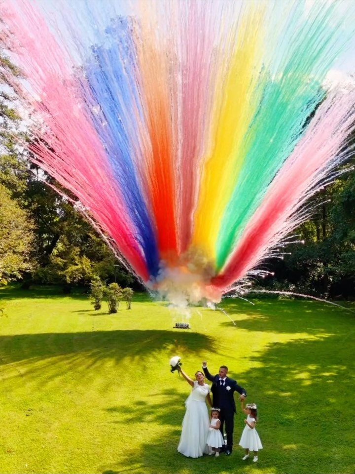 A wedding party stands on a lawn beneath a massive rainbow-colored smoke bomb explosion.
