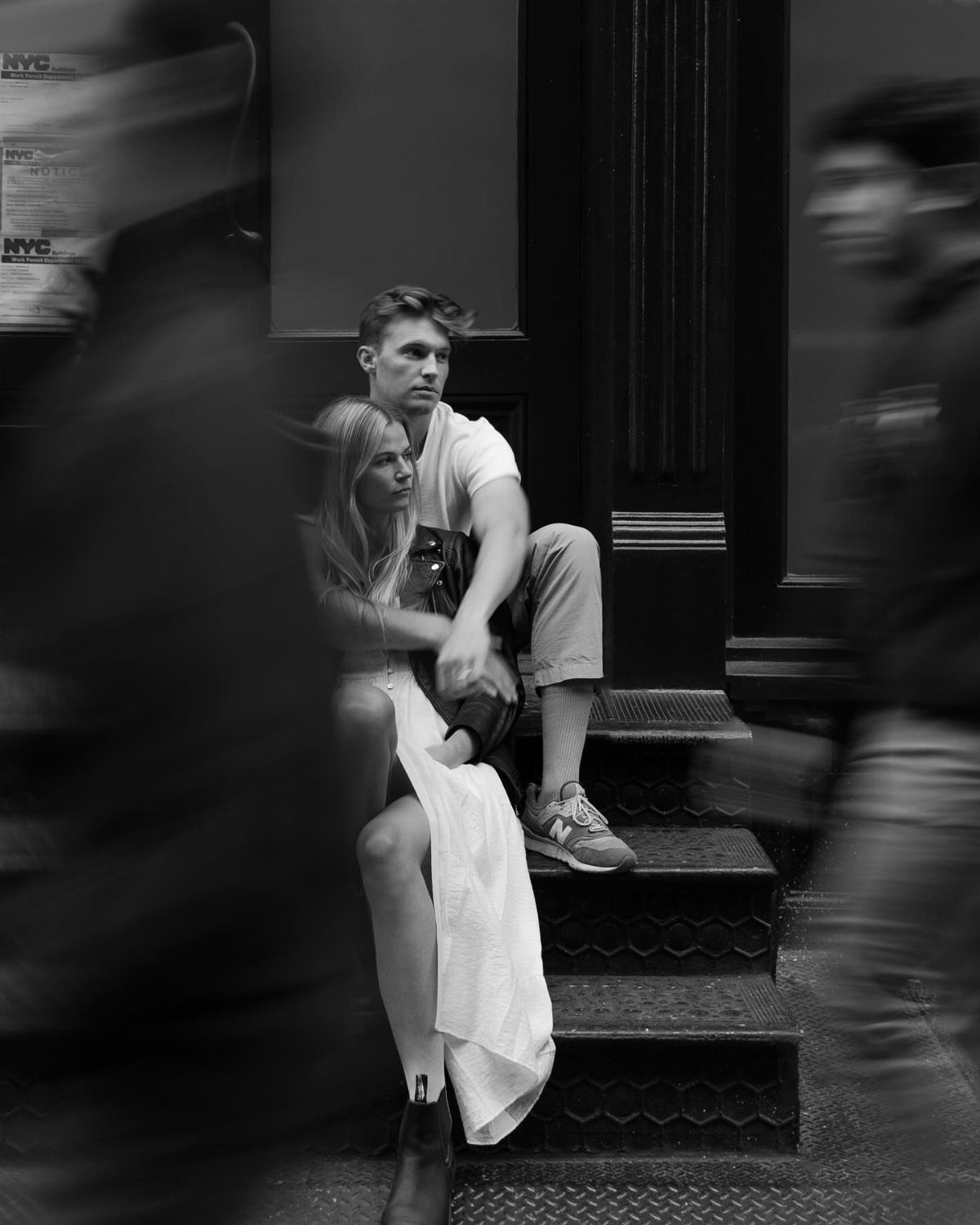 A young couple sits on city stairs while blurred pedestrians walk past them in black and white.