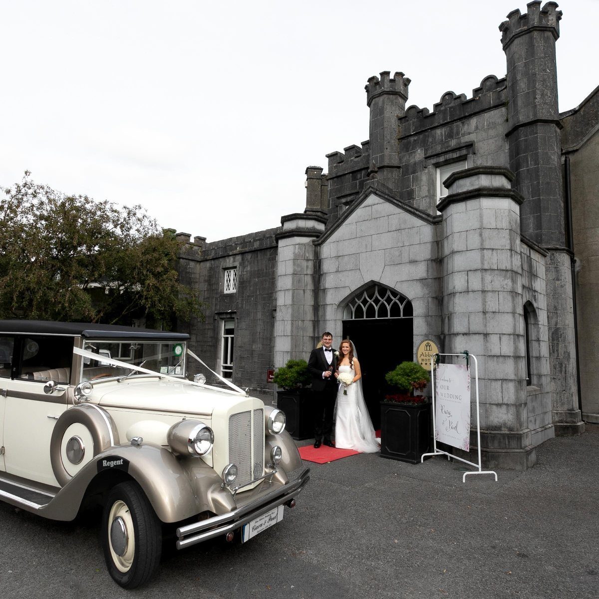 A bride and groom standing by a vintage car in front of a stone castle.