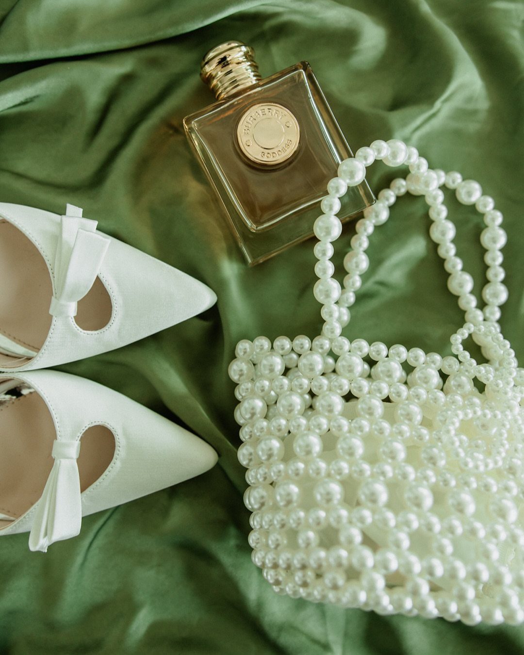 White heels, a pearl handbag, and a perfume bottle resting on green satin fabric.
