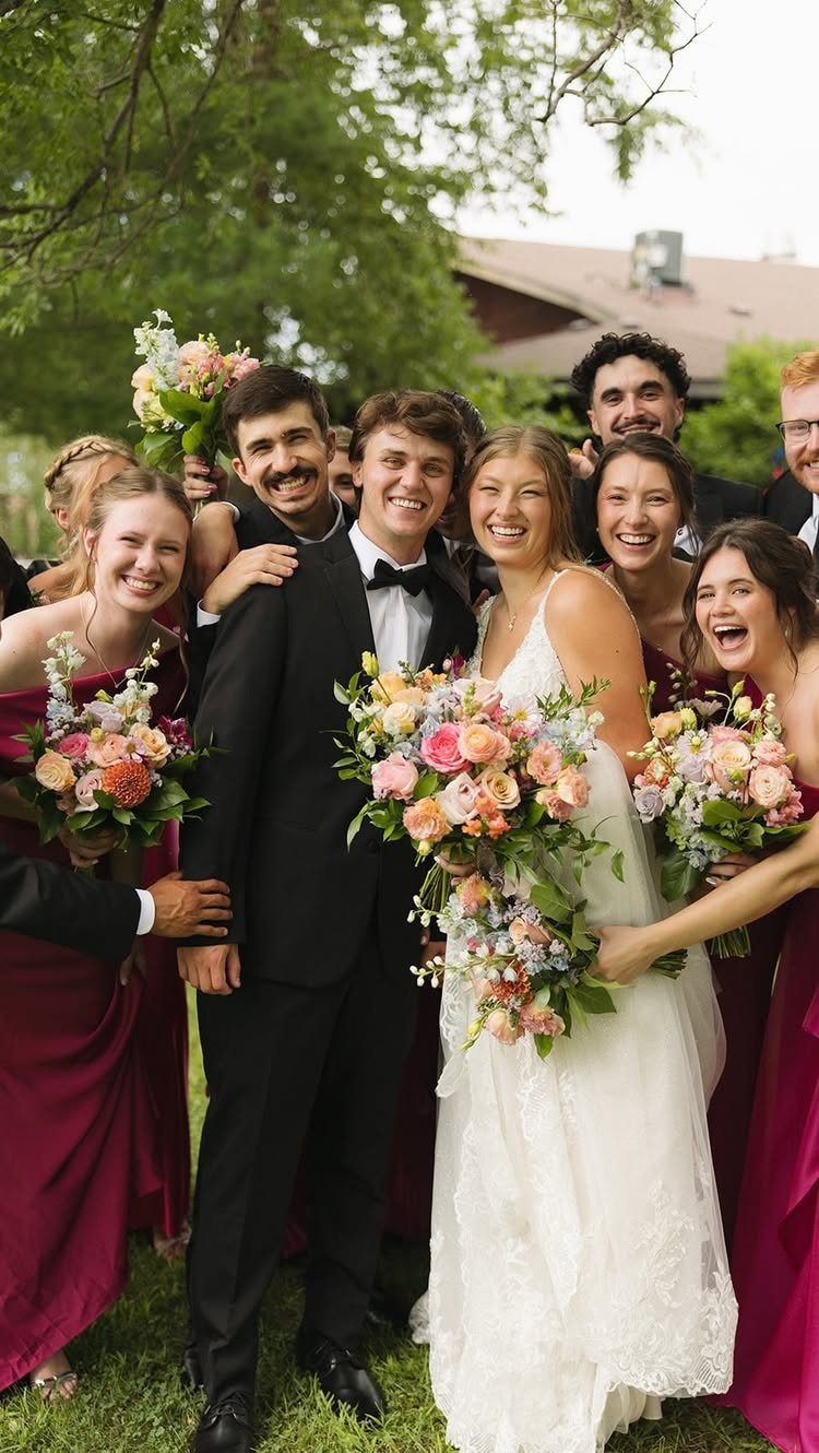 A smiling wedding party posing together outdoors with colorful floral bouquets and burgundy dresses.