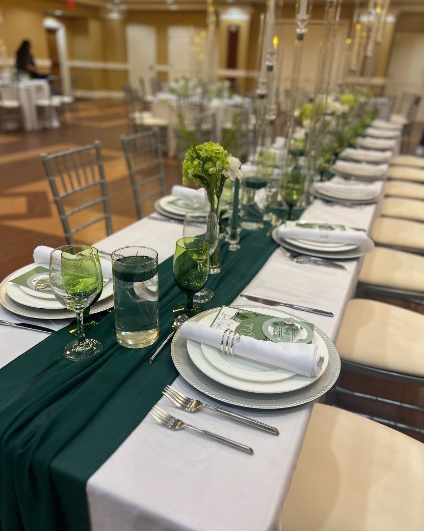 11 Wedding Party Table Decor Ideas for Any Venue Style 3 A long banquet table decorated with a dark green fabric runner, white plates, and green glassware.