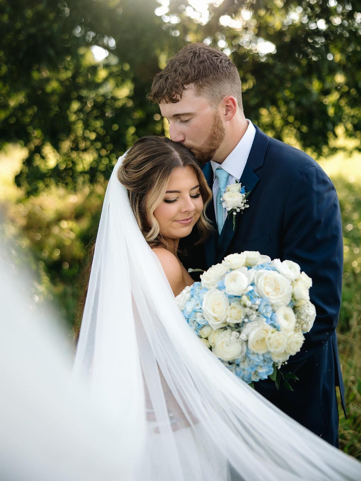 13 Wedding Photo Ideas for Your Shot List 2 A groom kissing the forehead of a bride holding a blue and white rose bouquet.