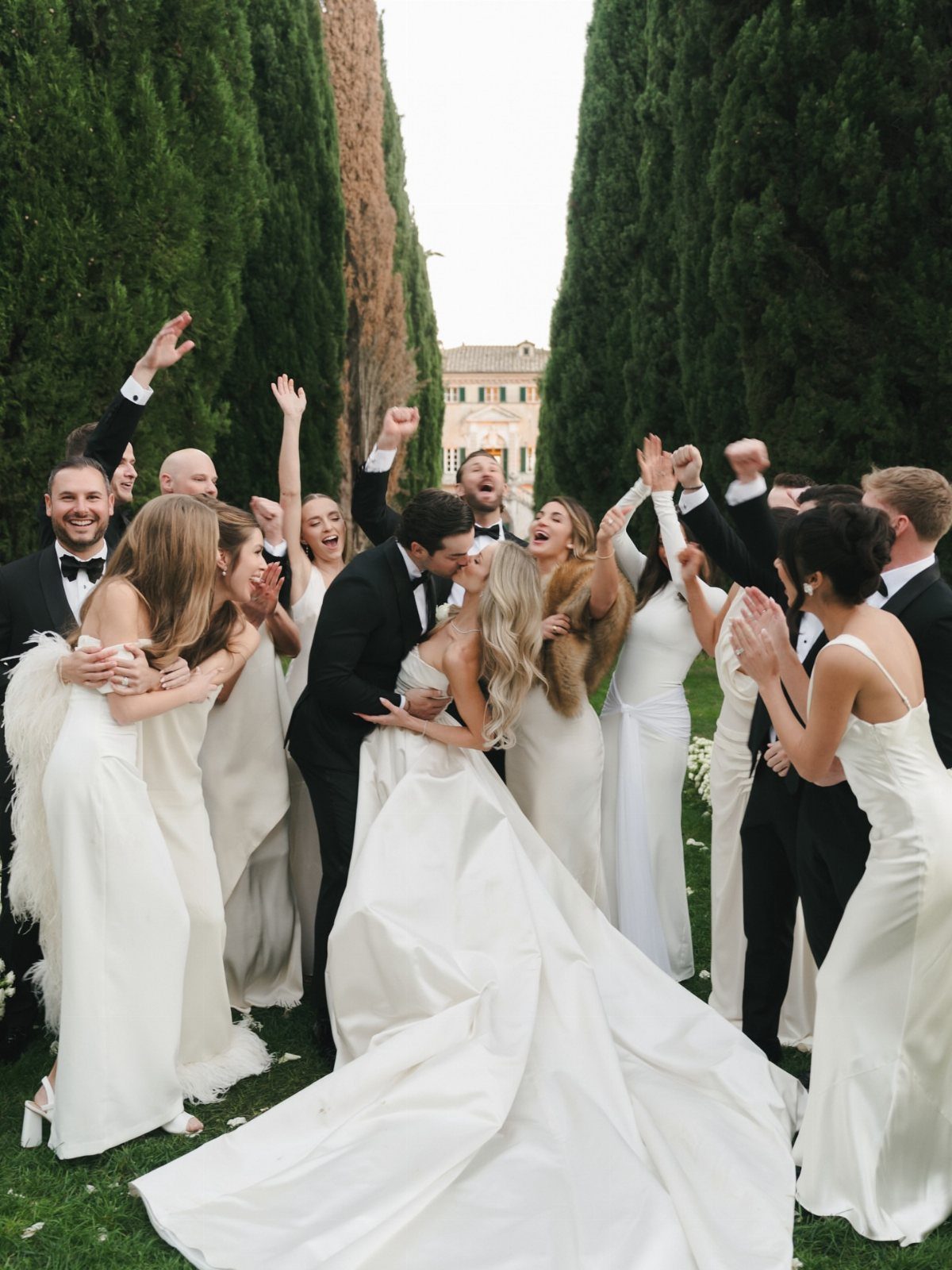 13 Wedding Photo Ideas for Your Shot List 8 Wedding party members in formal attire cheering and celebrating around a kissing bride and groom outdoors.