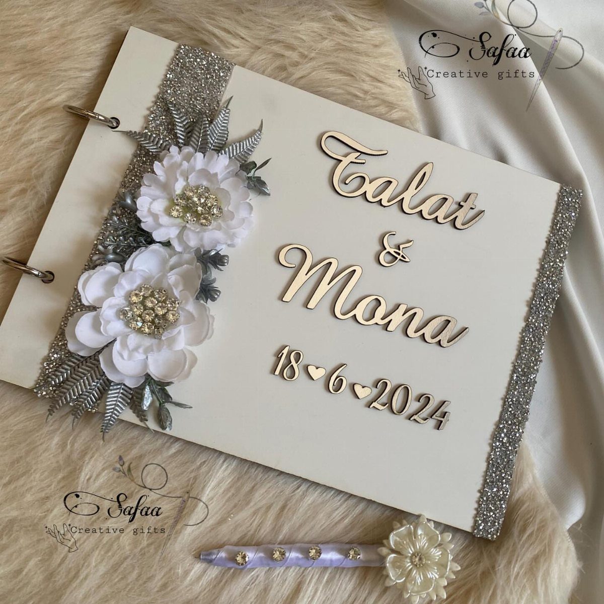 A white wedding guest book decorated with silver glitter, white flowers, and gold names.