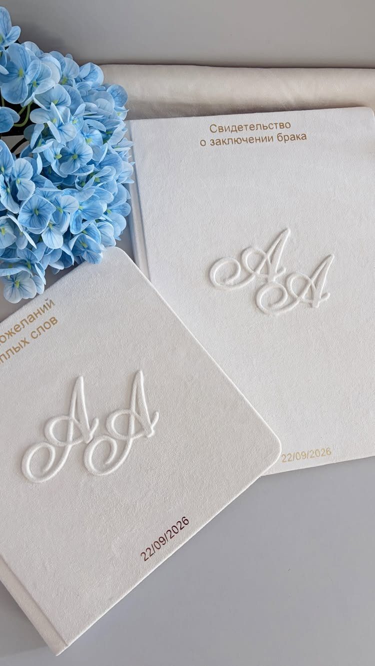 Two white embossed wedding books next to blue hydrangea flowers on a light surface.