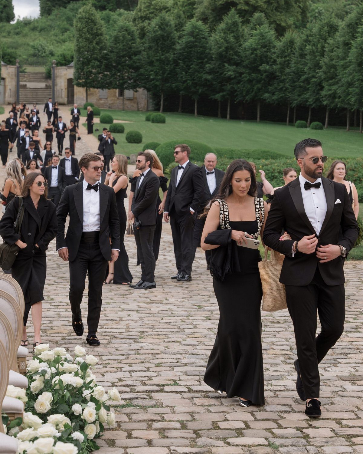 Wedding guests in black formal attire walking along a cobblestone path in an outdoor garden setting.