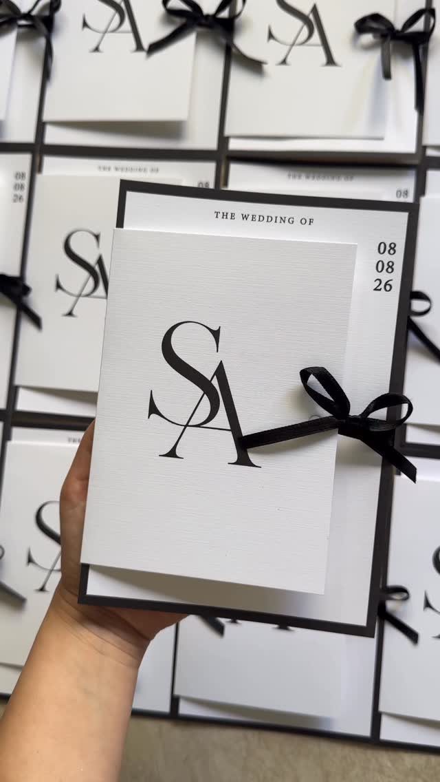 A hand holding a white and black wedding invitation suite with a monogram and ribbon.