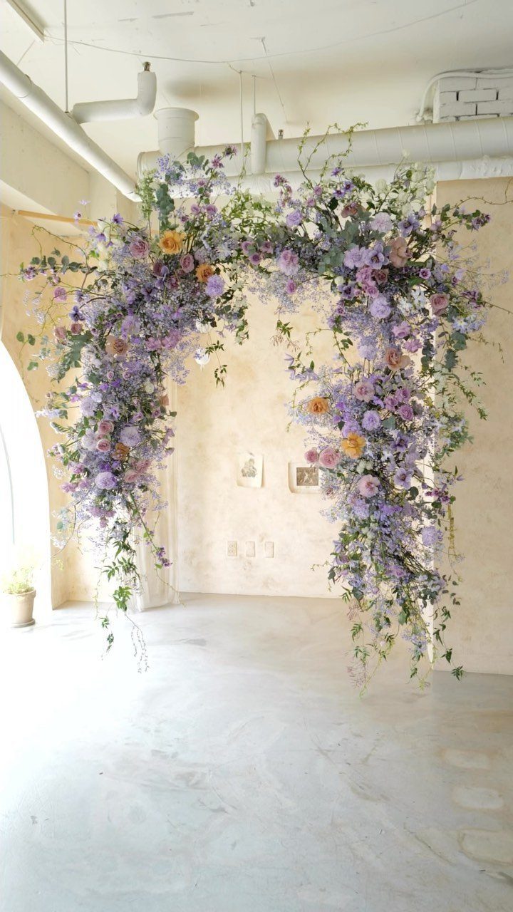 20 Spring Wedding Theme Ideas for 2026 7 A large floral archway featuring purple and peach flowers against a light wall.