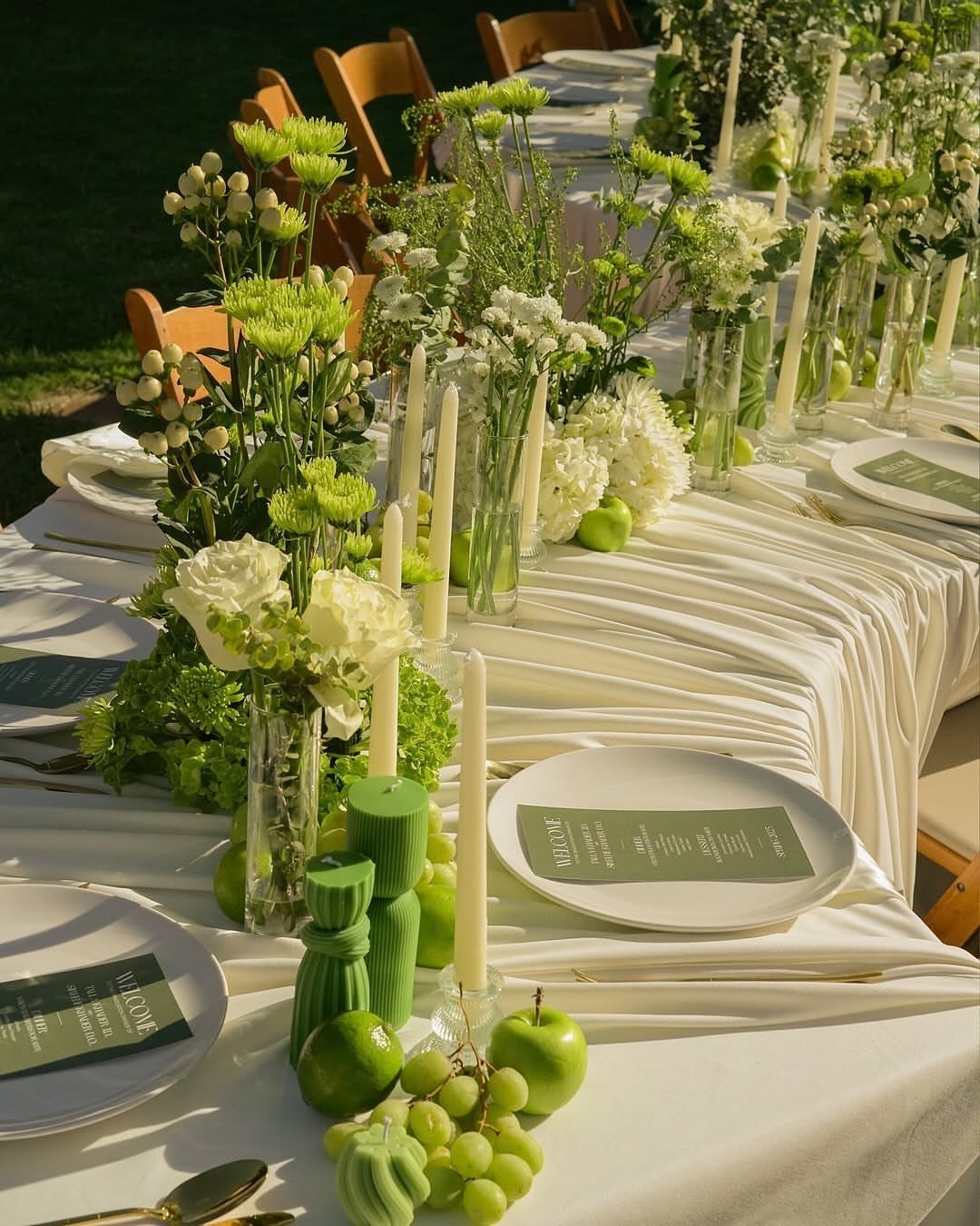 20 Spring Wedding Theme Ideas for 2026 10 A long wedding table decorated with white flowers, tall candles, and green fruit centerpieces.