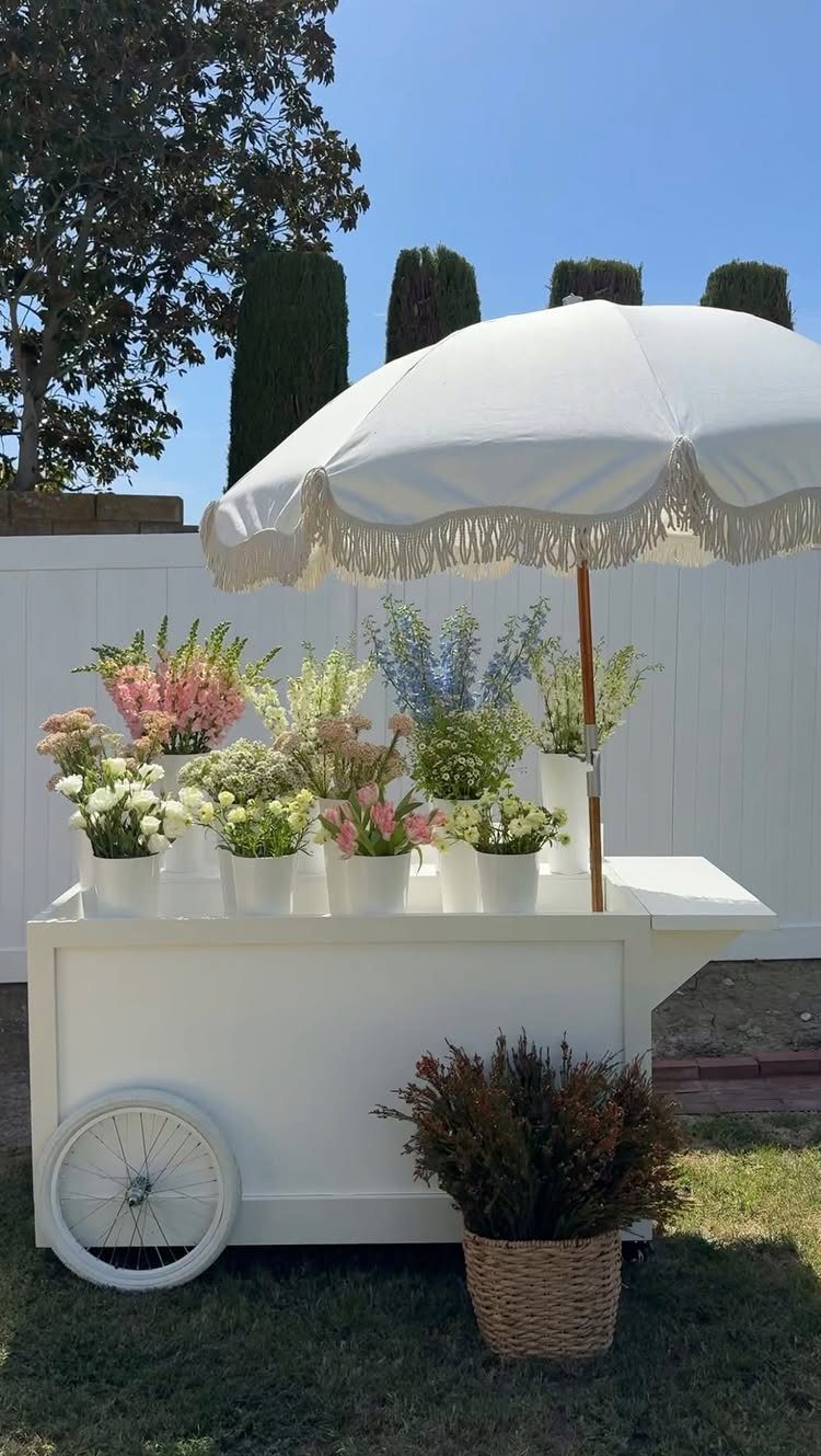 20 Spring Wedding Theme Ideas for 2026 9 A white flower cart with various colorful blooms under a white fringed umbrella outdoors.