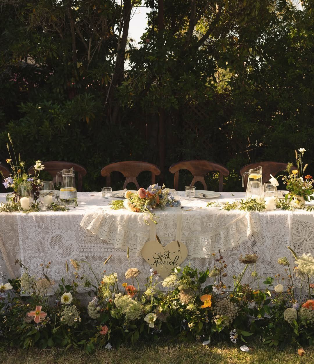 20 Spring Wedding Theme Ideas for 2026 14 A long wedding table with lace tablecloth, wildflowers, and a Just Married sign outdoors.
