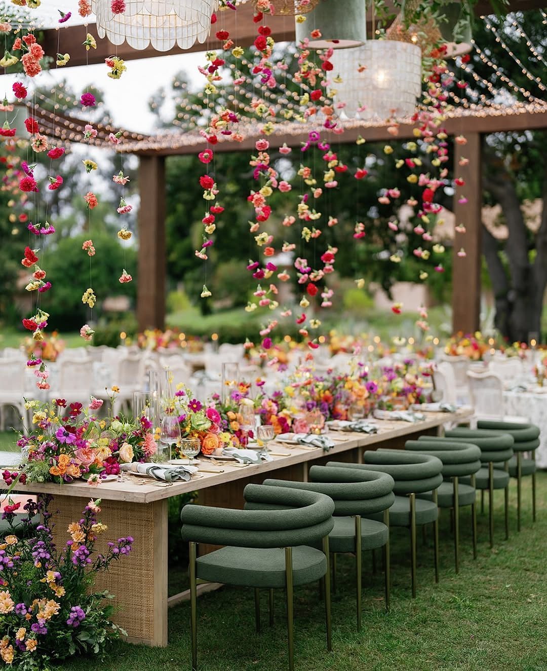 20 Spring Wedding Theme Ideas for 2026 3 Long wooden dining table decorated with colorful flowers under a pergola in an outdoor garden setting.