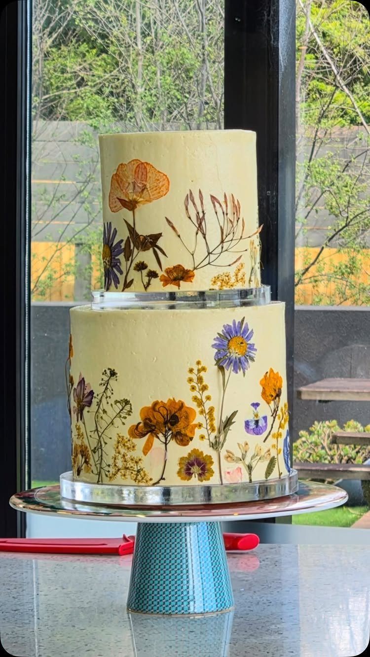 20 Spring Wedding Theme Ideas for 2026 2 Two-tier yellow cake decorated with colorful pressed wildflowers on a silver stand.