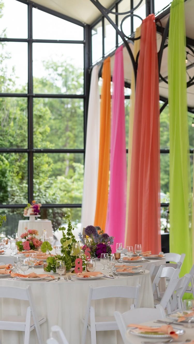 20 Spring Wedding Theme Ideas for 2026 15 Wedding reception tables with colorful floral centerpieces and bright hanging fabric drapes by large windows.