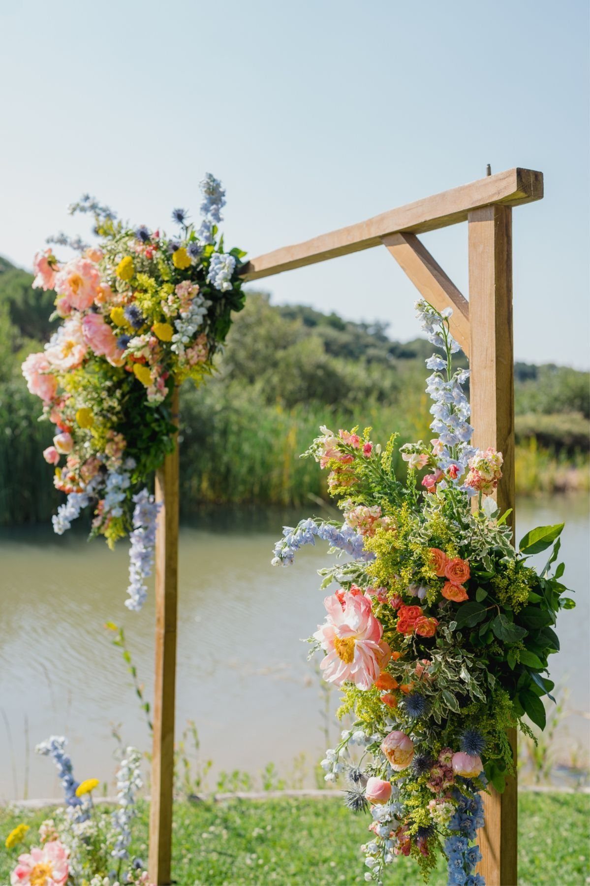A wooden wedding arch adorned with colorful wildflower arrangements standing by a lake.