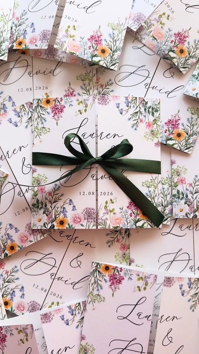 Scattered wedding invitations with colorful wildflower illustrations and a dark green satin bow.