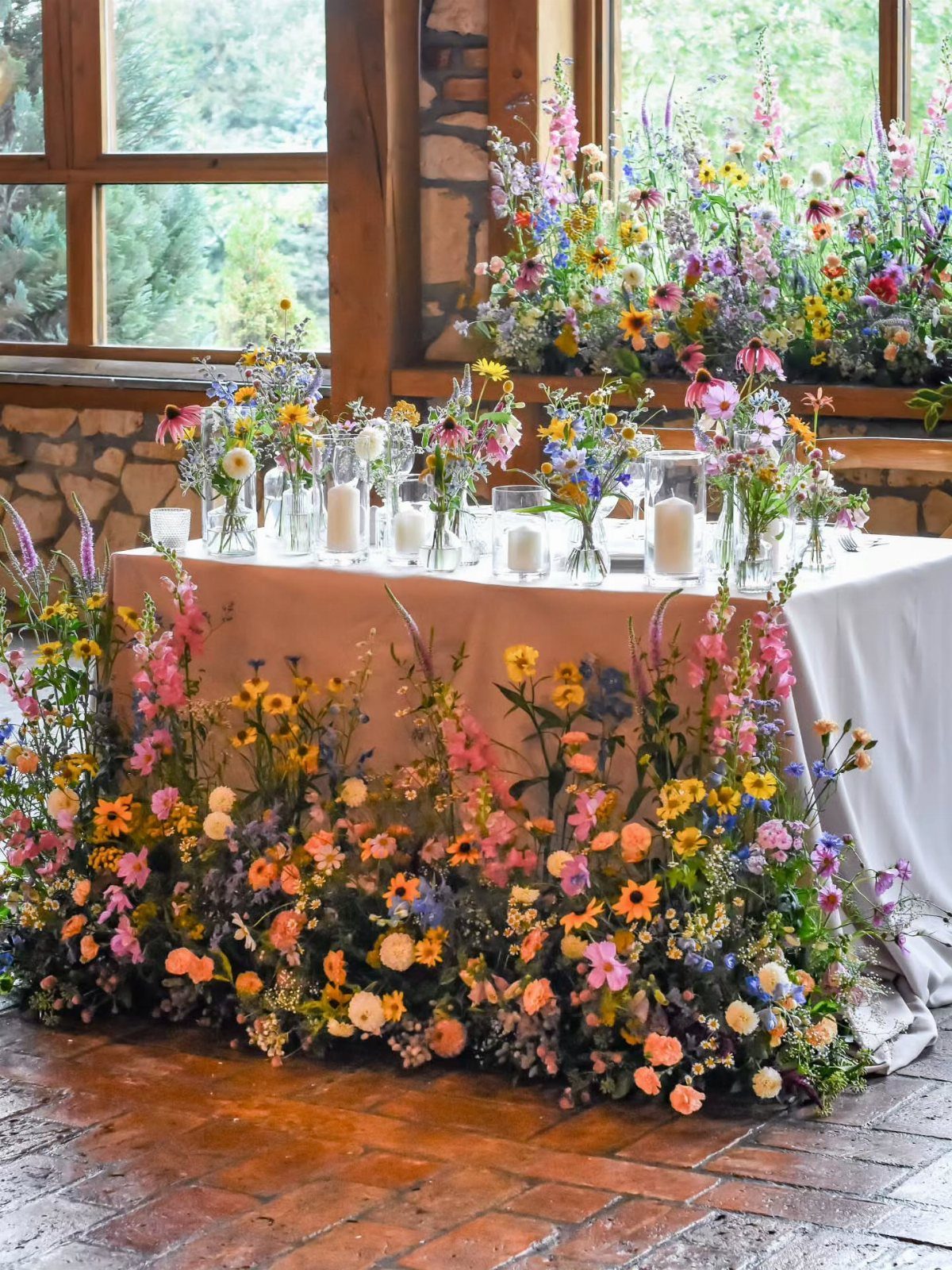 Colorful wildflowers in glass vases arranged on a long table with floral arrangements at the base.