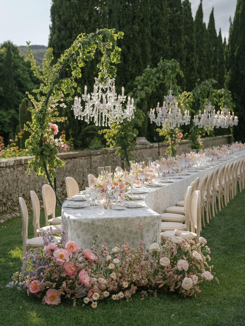 A long wedding table decorated with white linens, glassware, and pink wildflowers under hanging crystal chandeliers.