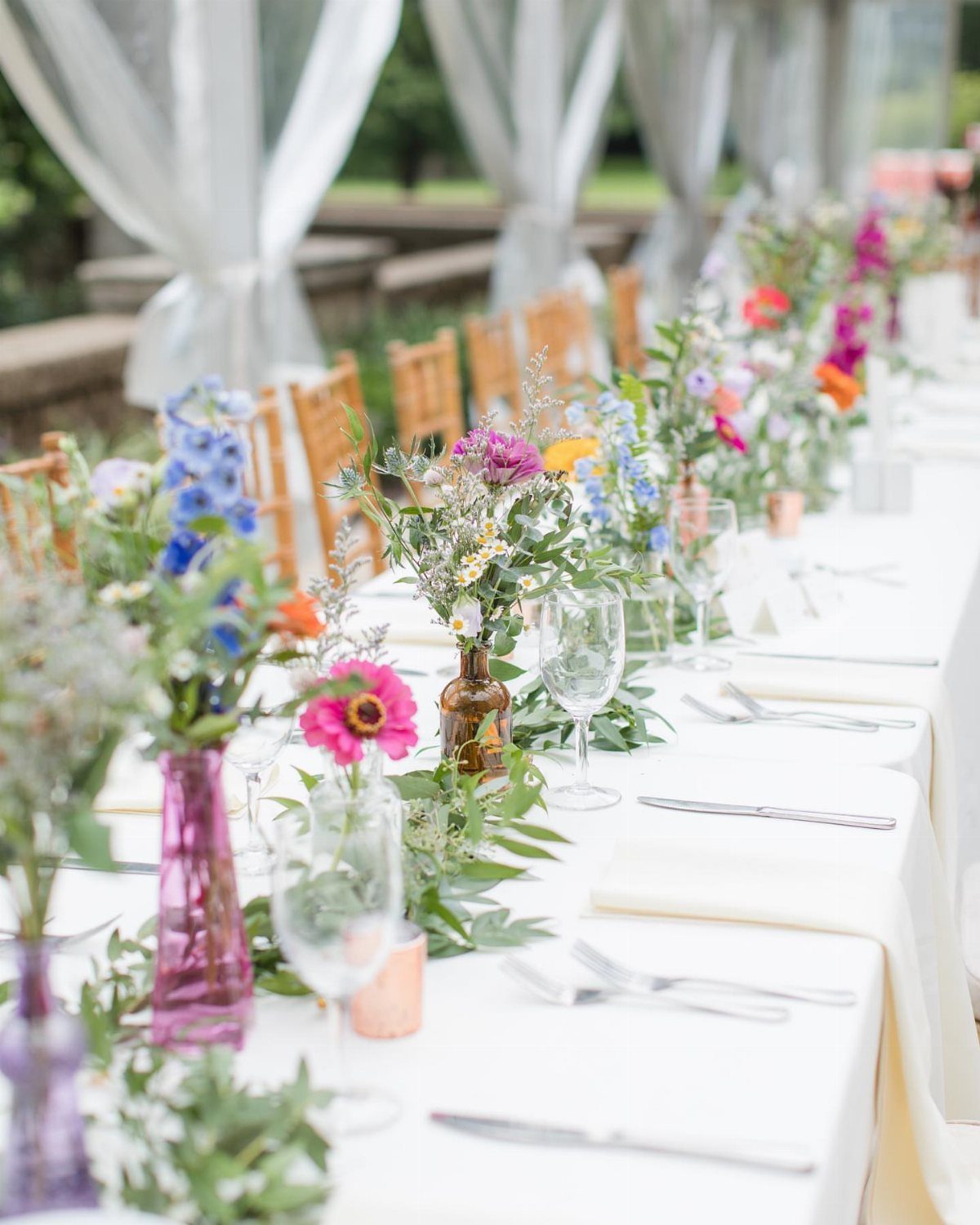 Colorful wildflowers in small glass vases arranged along a long white wedding reception table.