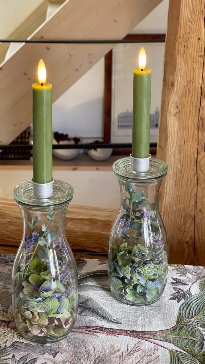 Two glass bottle candleholders filled with wildflowers and greenery, topped with lit green candles.